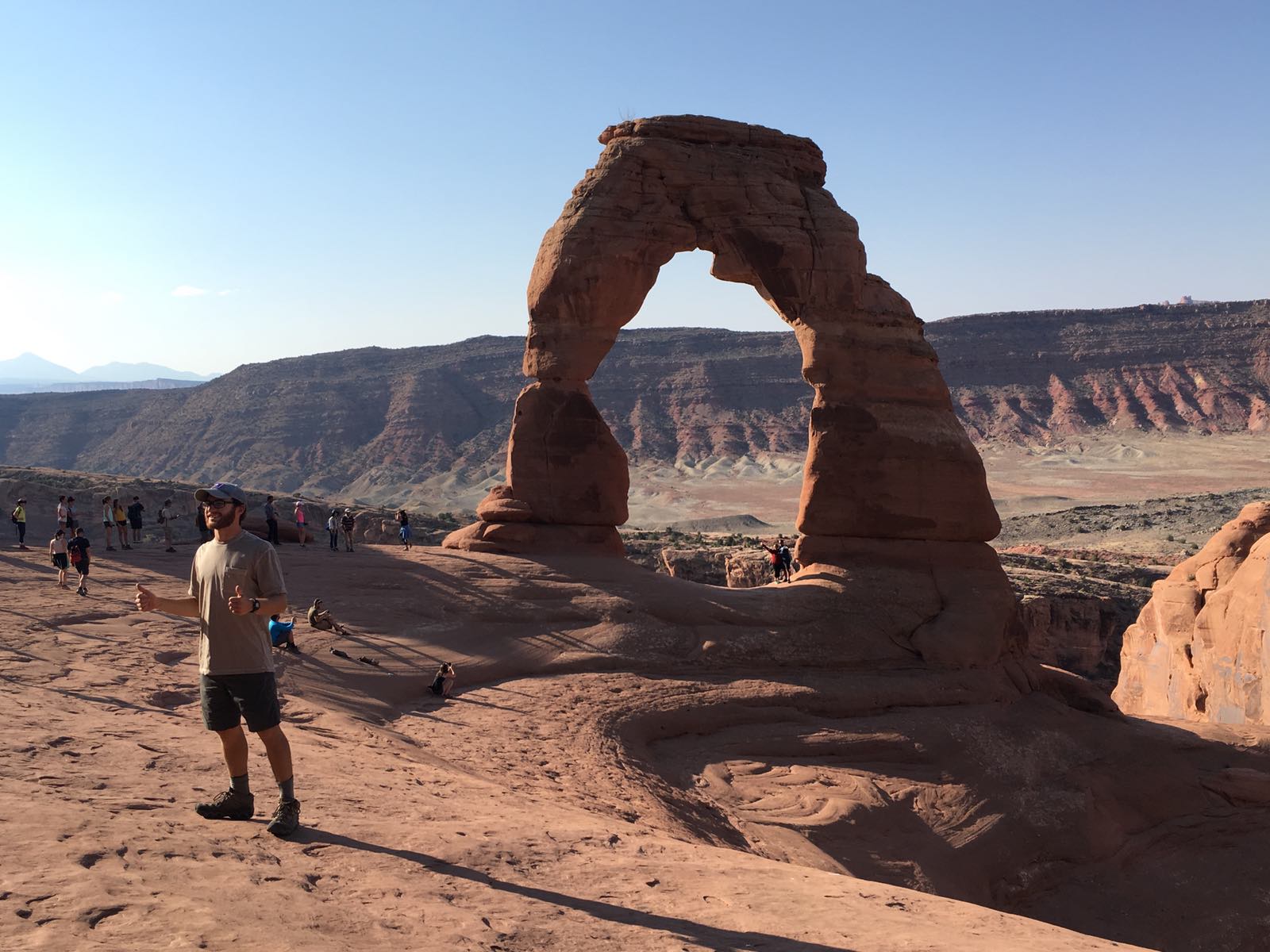 Delicate arch in background, in foreground a young man gives two thumbs up.
